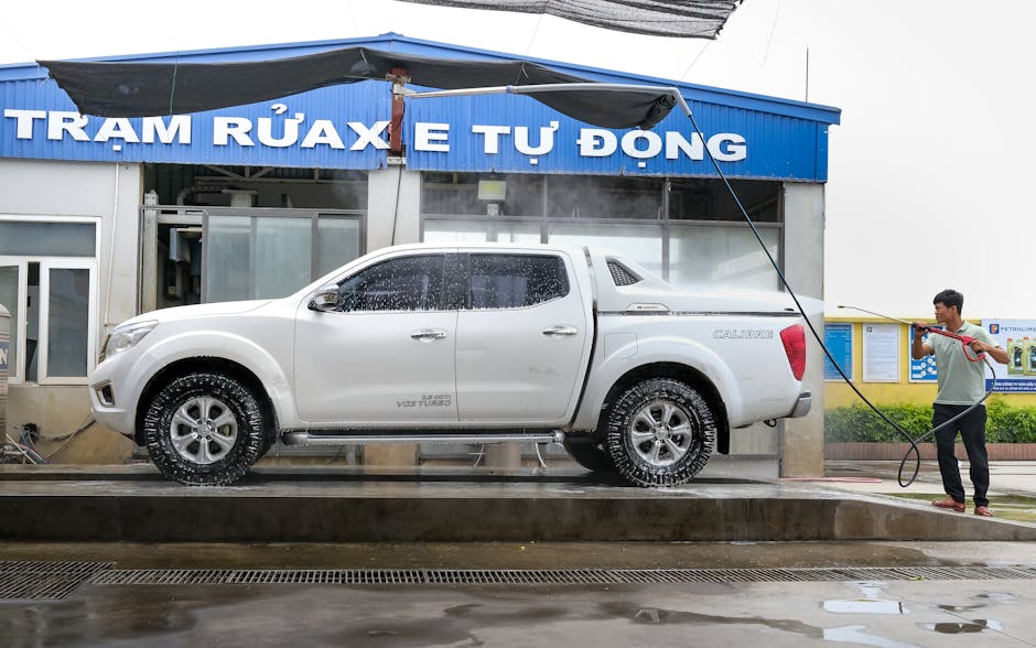 Adult using pressure washer to clean a white pickup truck at an outdoor car wash facility.